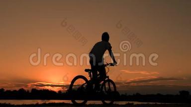 一个穿T恤的青少年坐在自行车上，在黎明的清晨骑着自行车穿过河流或湖泊。 骑自行车的人的剪影